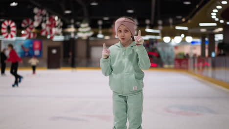 a close view of a child in a mint green outfit and pink headband dancing joyfully on an ice rink, the background shows other people skating