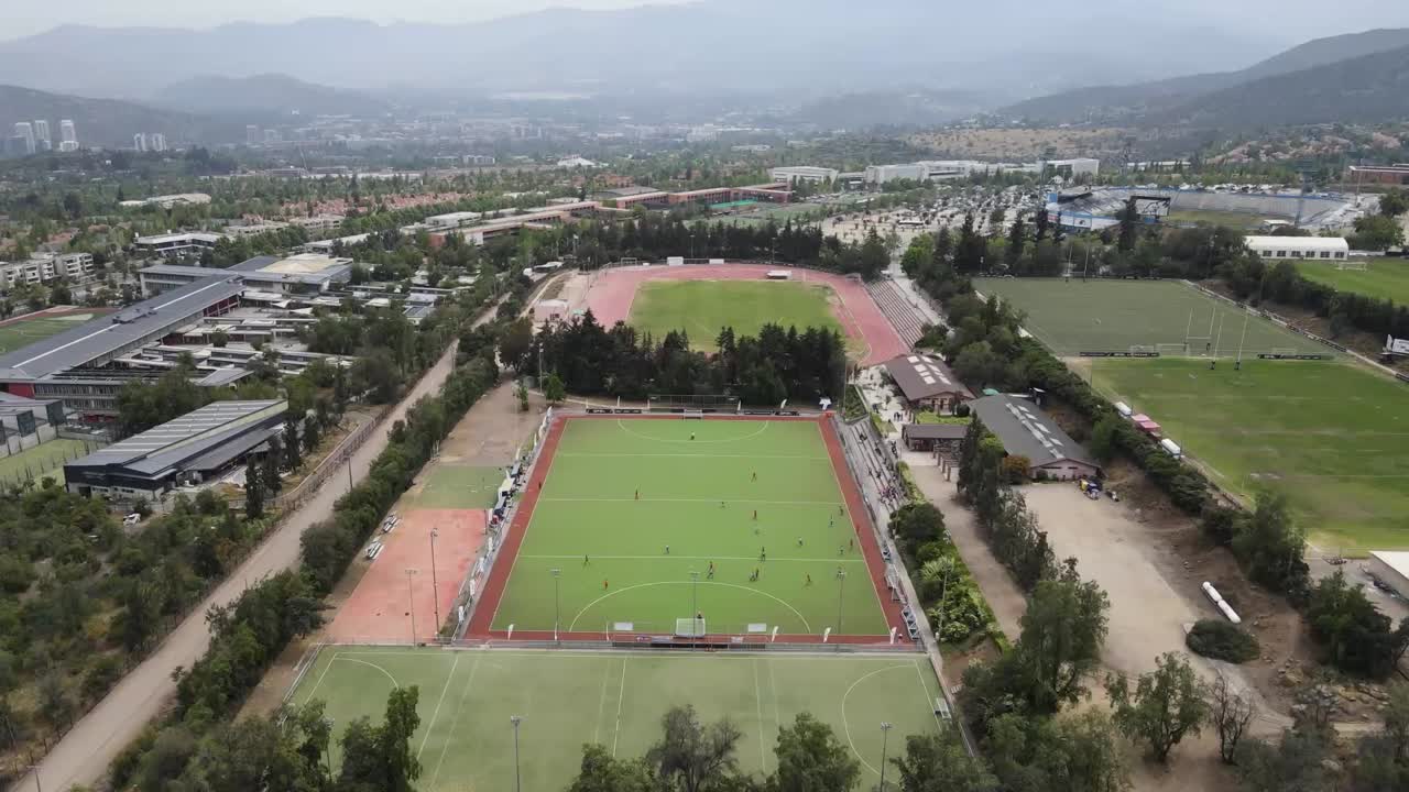 Field hockey players competing intensely on verdant playing field, urban cityscape surrounding athletic action and highlighting team dynamics against architectural backdrop