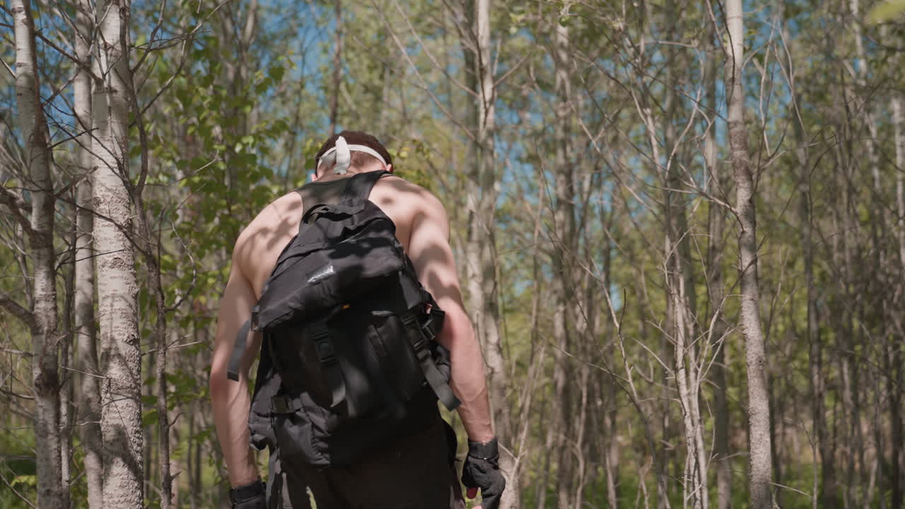 Back view of adventurer carrying black pack through sunlit dense forest stepping over fallen branches in worn jungle boots on leafy trail under vibrant green canopy in midday light