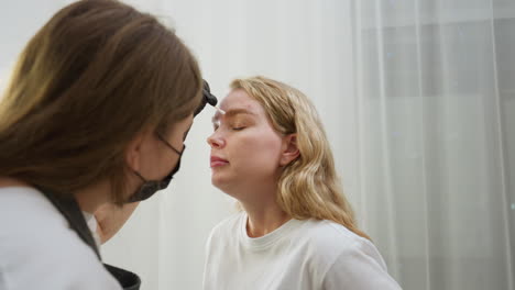 beautician in mask adjusts client eyebrow using cotton wool while standing close in well lit beauty studio with soft white curtain and ring light visible and client remains relaxed with eyes closed