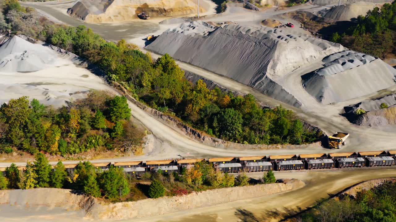 Aerial of dump truck driving in quarry with gravel piles, conveyor belts and freight train, slow motion tracking left