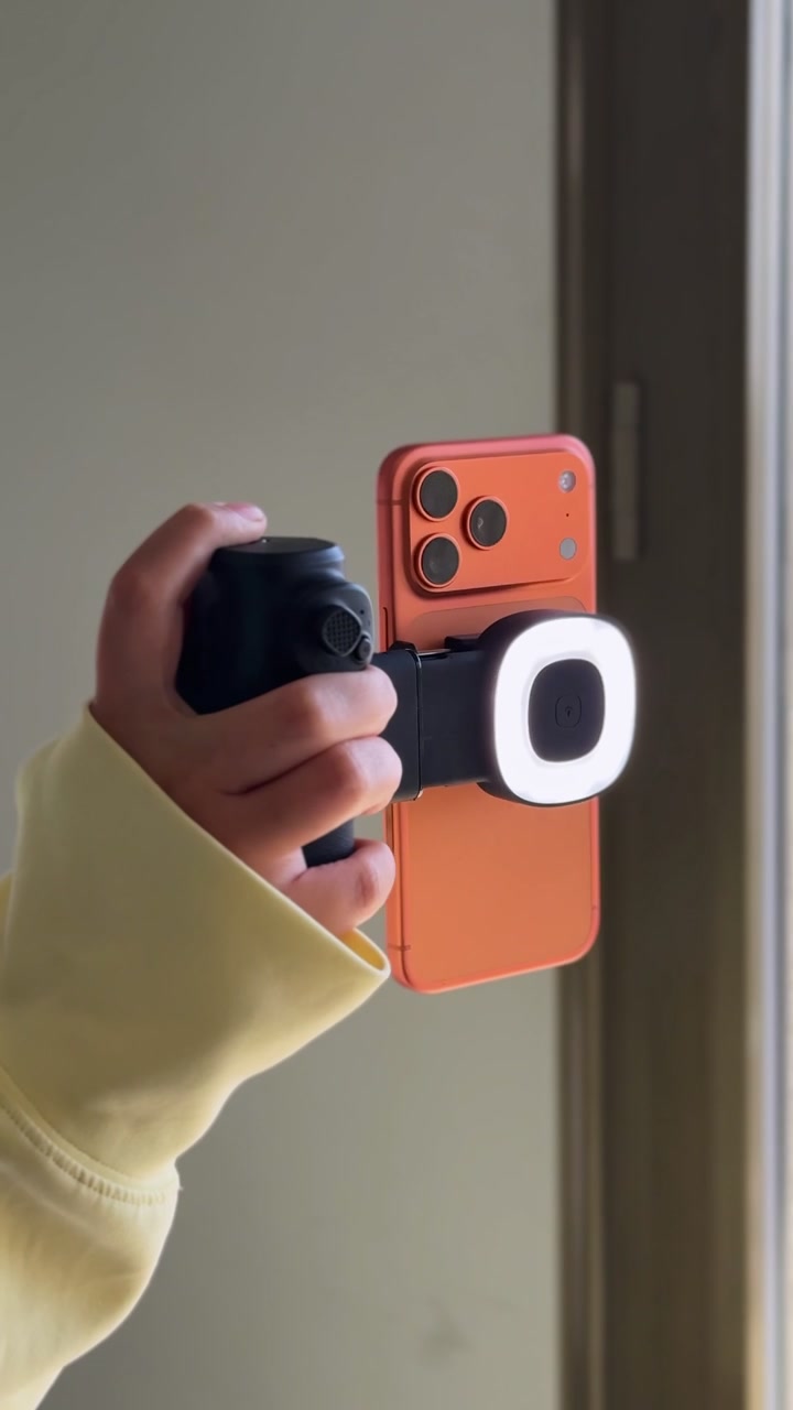 Close-up de uma mão segurando um smartphone laranja em um rig de filmagem portátil com um ring light LED iluminado, preparado para gravação de vídeo vertical em ambiente interno.