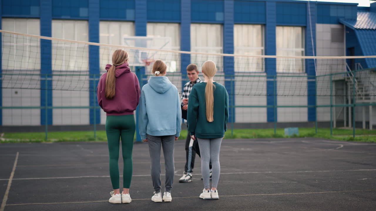 젊은 배구 선수들이 야외 코트, 네트, 파란색 건물, 그리고 배경에 울타리로 둘러싸인 구역에서 훈련하는 동안 지침을 주는 코치의 말을 주의 깊게 듣고 있다.