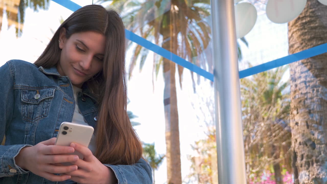 mujer joven sonriente enviando mensajes de texto por teléfono sentada en la parada de autobús en un día soleado