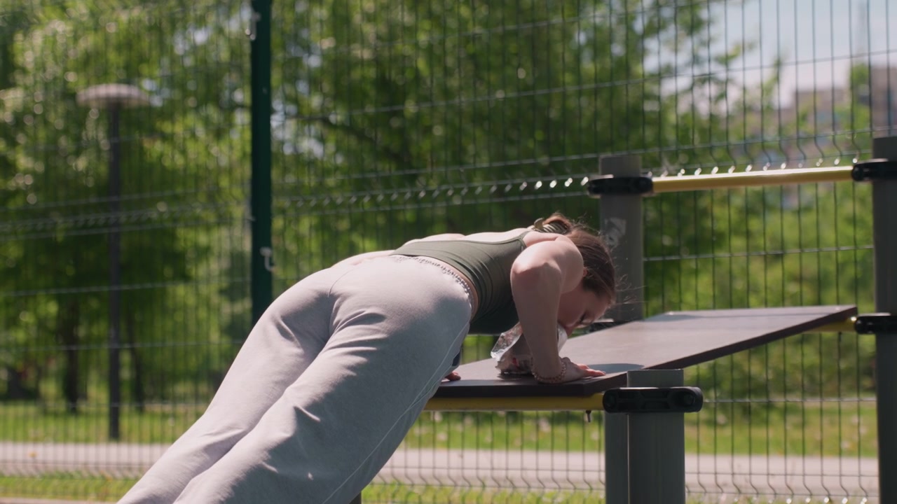 Close up of young woman performing push ups on outdoor fitness equipment at park with green trees and fence in background. Woman wears dark sports bra and light gray sweatpants maintaining