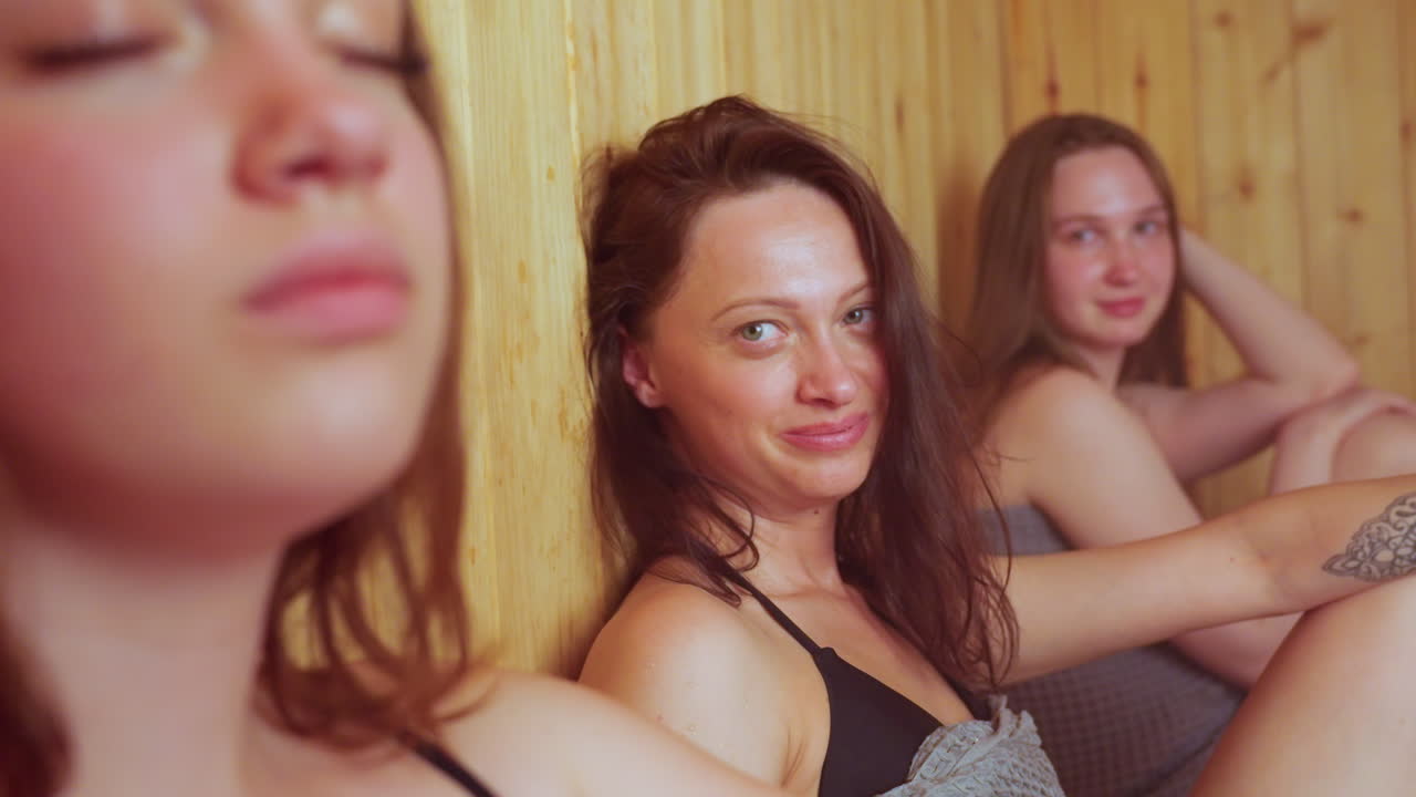 close up of woman fanning herself inside warm sauna while two women seated nearby glance toward her with gentle smiles, surrounded by smooth wooden walls and soft steamy atmosphere