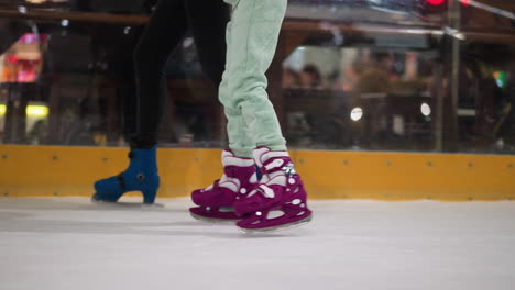 vista en primer plano de dos personas patinando en una pista de hielo, una con patines púrpuras con pantalones verdes menta, y la otra con patines azules con pantalóns negros, el fondo muestra una vista borrosa de personas