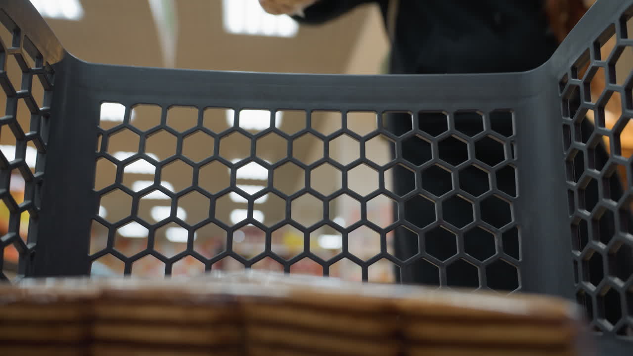 vista de una persona moviendo un carrito de compras con un paquete de galletas adentro mientras una dama con un abrigo negro y una mochila coloca un trozo de pan en el carrito