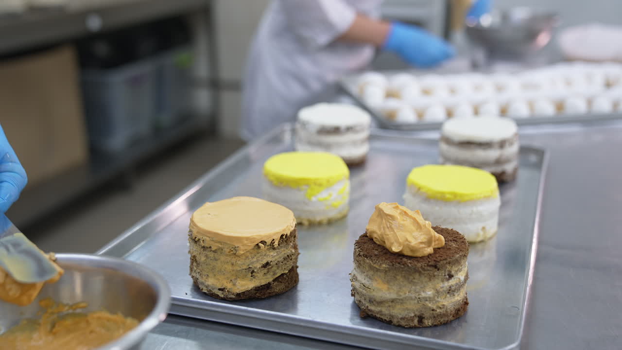 Metal spatula is used to spread cream over the cake surface. Varied cakes are placed on metal tray for decoration. Close up.