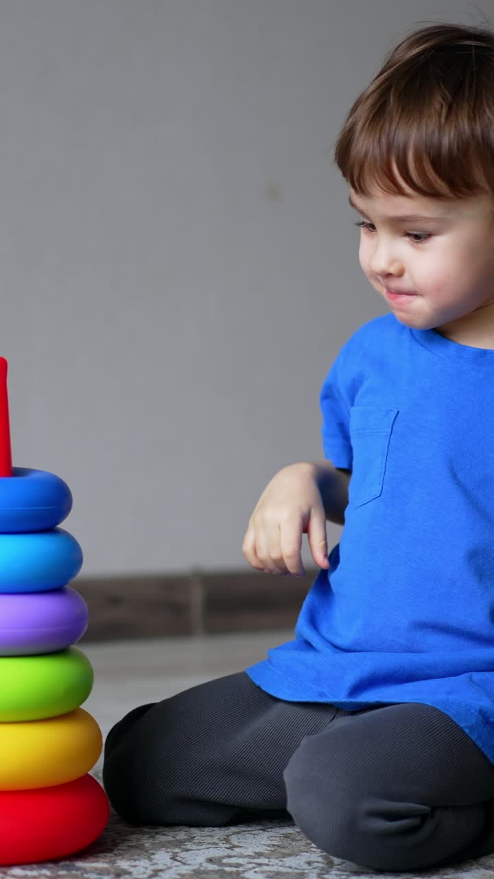 Un bambino caucasico sorridente gioca con una piramide giocattolo. Il bambino mette gli anelli sulla base allegramente e poi ricomincia. Video verticale.