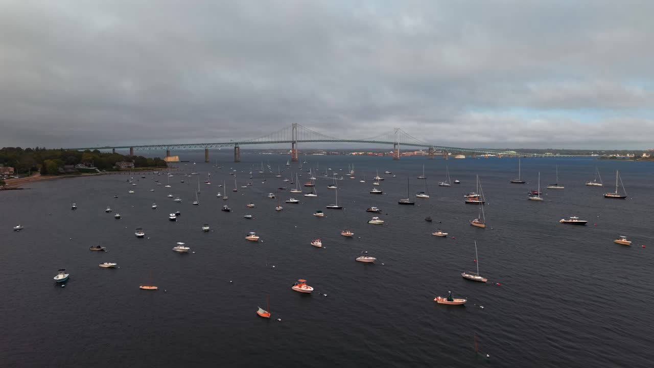 An aerial view of a marina, south of the Claiborne Pell-Newport Bridge on a cloudy day in Jamestown, Rhode Island. The drone camera boom up over the water showing the anchored boats and the bridge