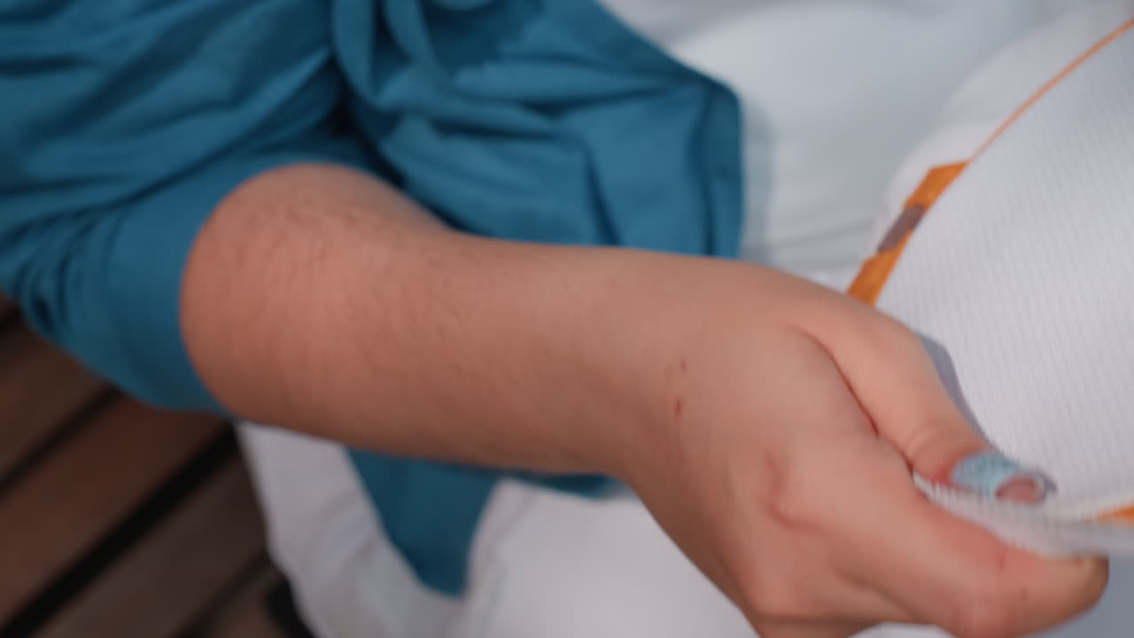 Nærbillede af kvinde, der vifte sig med stof i varmt sollys, blød håndbevægelse, lyseblå negle synlige, blå ærme over hvidt outfit, gestus, der udtrykker varmelettelse og afslapning