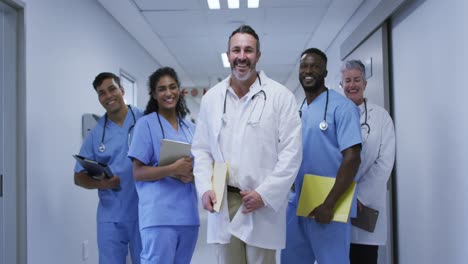 Diverse-group-of-smiling-male-and-female-doctors-in-hospital-corridor-wearing-scrubs-and-lab-coats