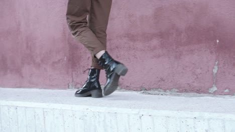 close-up-of-a-woman-walking-forward-wearing-shiny-black-boots-and-brown-pants-at-an-urban-set-up-with-a-red-wall-as-a-background