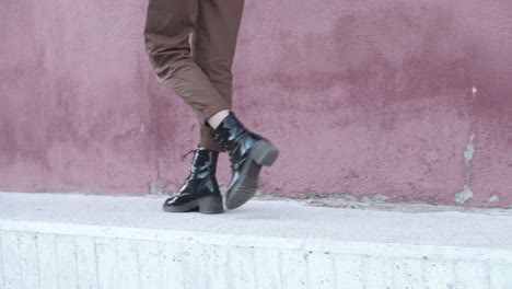 El-Primer-Plano-De-Una-Mujer-Que-Camina-Hacia-Adelante-Con-Botas-Negras-Brillantes-Y-Pantalones-Marrones-En-Un-Entorno-Urbano-Con-Una-Pared-Roja-Como-Fondo