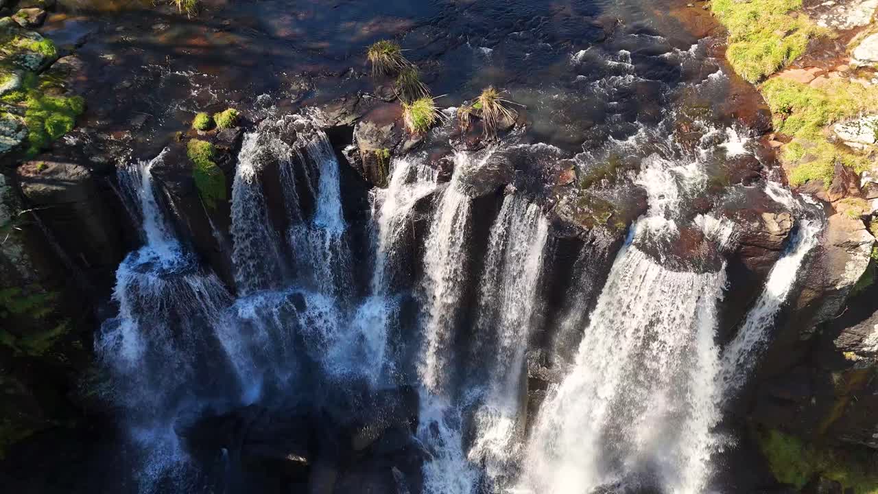 Drone footage captures cascading waterfalls at Ebor Falls, New South Wales, Australia. Bright daylight highlights lush greenery, rocky cliffs, and flowing water from a high, top-down perspective