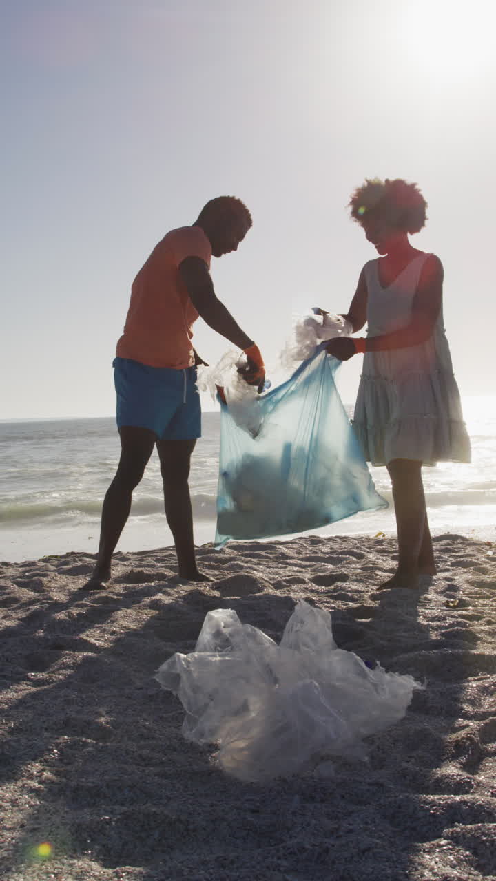 Video verticale di coppia afroamericana felice che differenzia i rifiuti su una spiaggia soleggiata