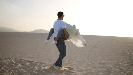 Escena-Perfecta-De-Pareja-Joven-En-El-Desierto-Vacío.-Hombre-Sosteniendo-A-Su-Mujer-En-Brazos-Y-Dándole-La-Vuelta,-Diviértete-En-El-Desierto-Loco-De-Amor,-Emociones-Y-Relaciones.-Montañas-En-El-Fondo