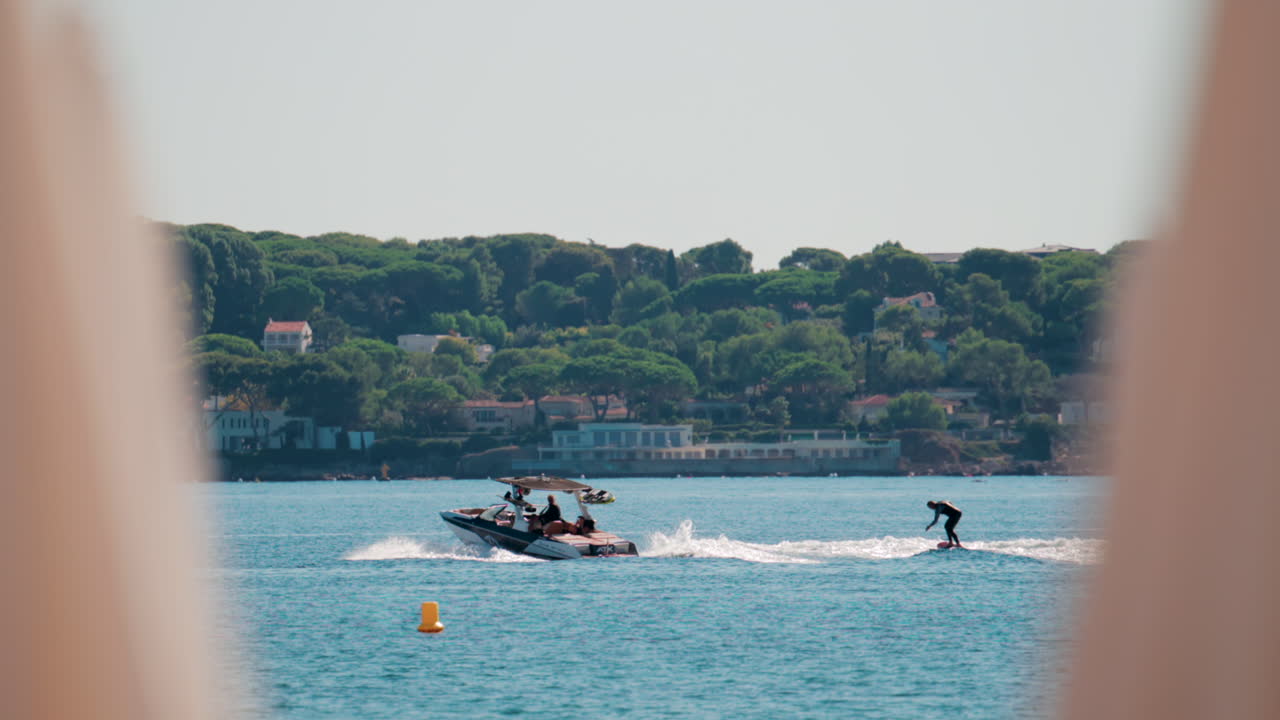 A wakeboarder glides over the waves behind a speedboat on a sunny coastal day, with scenic villas and pine covered hills in the background