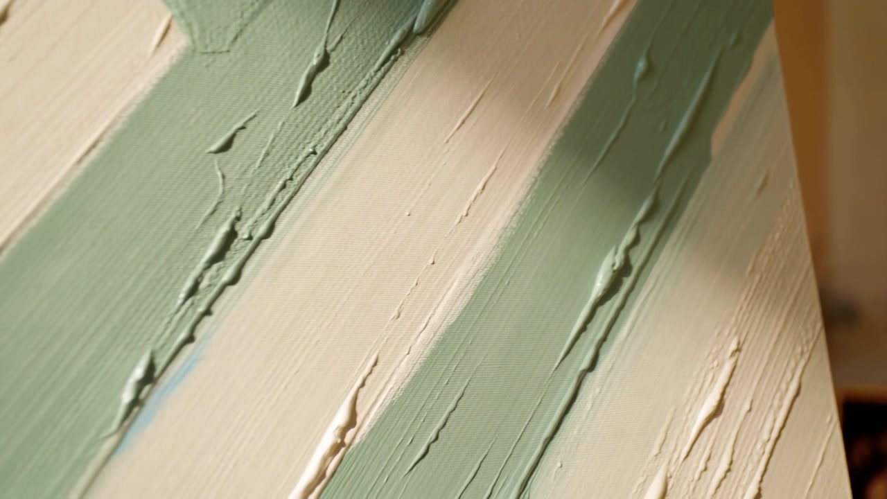 Primo piano obliquo di una tela con bande diagonali alternate di verde salvia e beige cremoso, realizzata con pittura a impasto spesso, sottili rilievi, luce calda e una raffinata texture superficiale astratta.