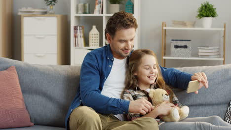 Adolescente-Linda-Chica-Rubia-Sentada-En-El-Sofá-Y-Abrazando-A-Su-Oso-De-Peluche-Mientras-Su-Padre-La-Abrazaba-Y-Mostraba-Algo-En-El-Dispositivo-Del-Teléfono-Inteligente