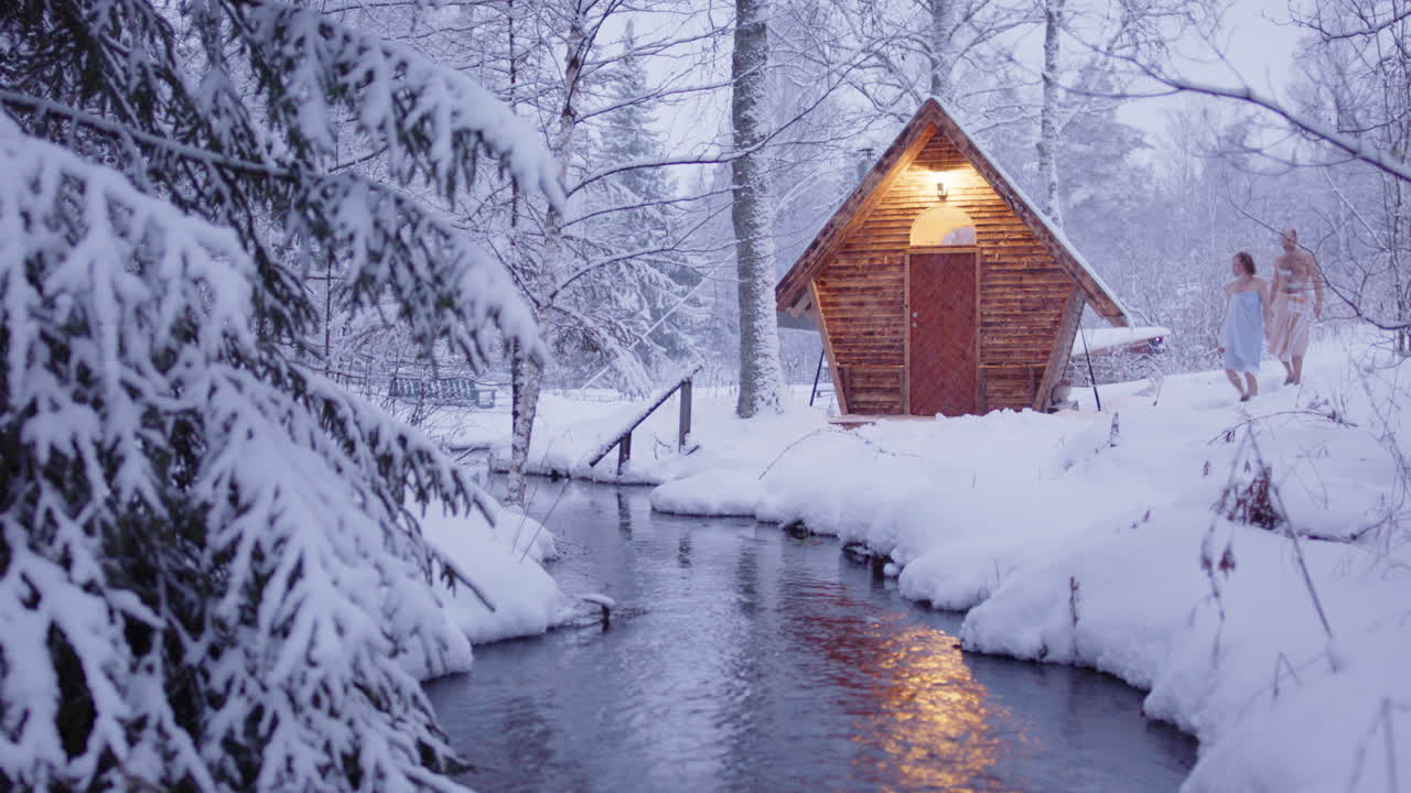 Couple in towels walk through snowy woods to wooden sauna next to stream, zoom