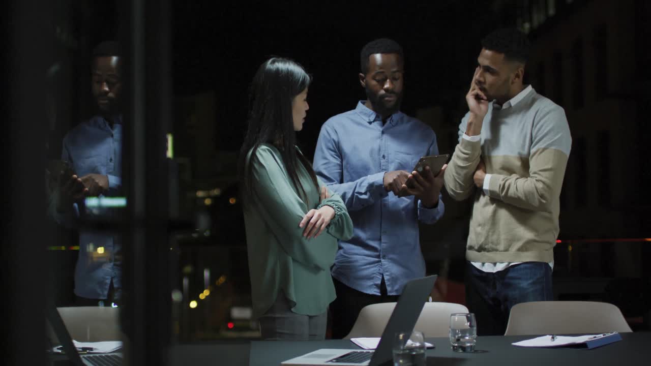 Video of three diverse colleagues talking standing and looking at tablet at night in office