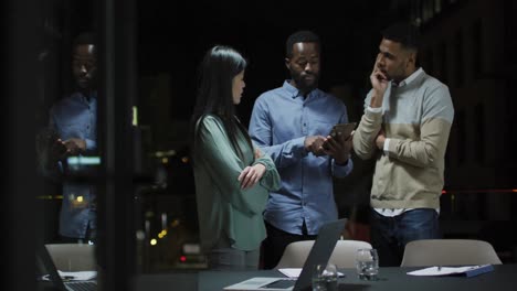 Video-of-three-diverse-colleagues-talking-standing-and-looking-at-tablet-at-night-in-office
