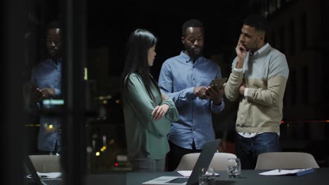 video of three diverse colleagues talking standing and looking at tablet at night in office