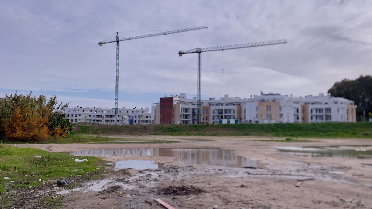 Nuovi edifici residenziali in costruzione con gru a torre e un'auto parcheggiata su terreno fangoso a Sanlucar de Barrameda, Spagna.