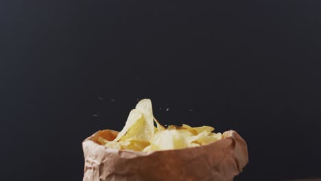 close up view of potato chips falling in a paper bag against black background