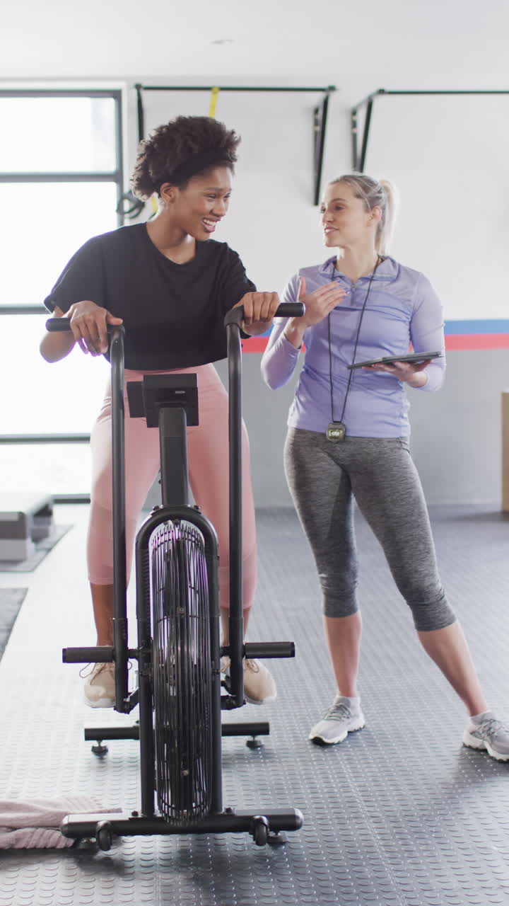 Vertical video of diverse, happy female fitness trainer and woman on exercise bike talking at gym