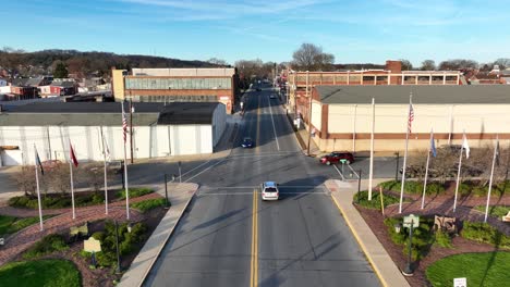 Drone-footage-of-a-city-park-with-American-flags,-benches-and-brick-walkways-next-to-an-industrial-district-in-the-springtime-at-sunset