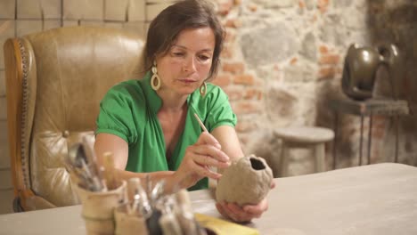 a front view of a beautiful female potter sitting in antique style armchair and glazing ceramics oval shaped sculpture using professional tools. artist working in studio.