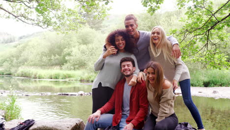 Five-young-adult-friends-take-a-break-and-pose-to-camera-standing-and-sitting-on-rocks-by-a-river-during-a-hike,-handheld