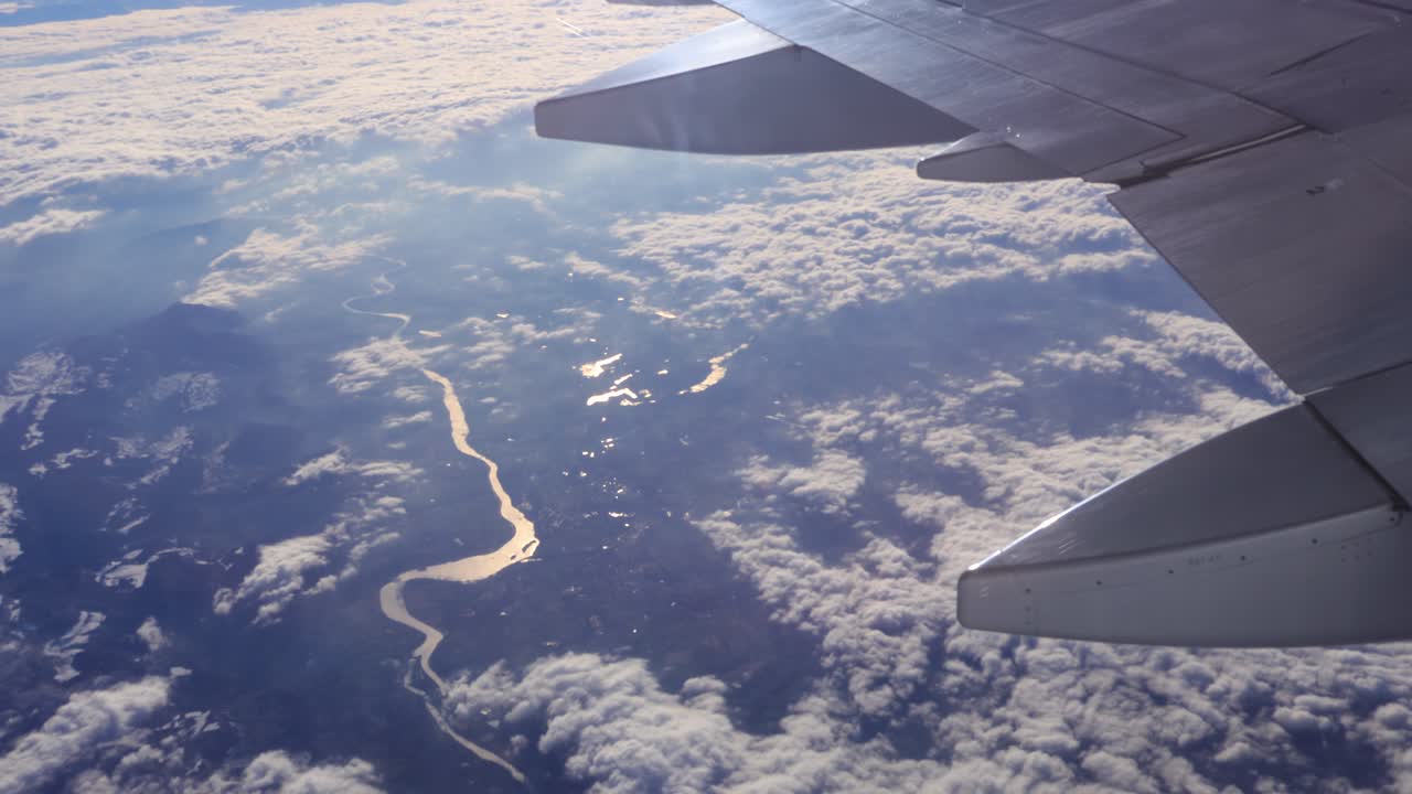 Udsigt fra et flyvindue, der viser en hvid vinge, der glider over spredte skyer og en snoet flod langt nede, med sollys der reflekteres fra vingen under en jævn flyvning.