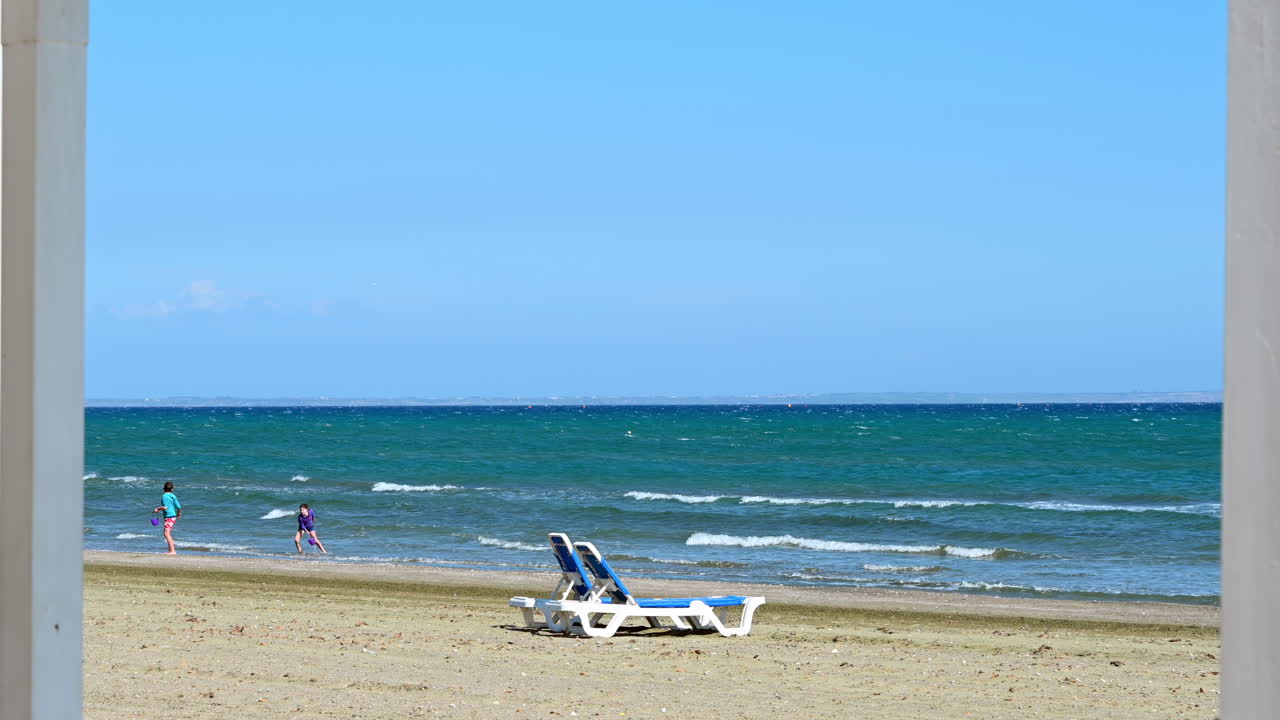 Limassol, Cyprus - April 16, 2019:Two empty blue sunbeds on the sandy shoreline with turquoise waves and a person walking along the beach