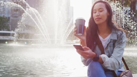 asian woman sitting next to fountain using smartphone and drinking takeaway coffee