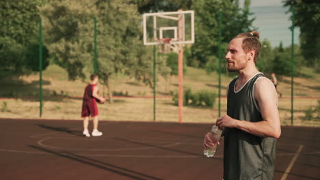 in foreground, a tired handsome blonde bearded basketball player taking a break and drinking water in an outdoor basketball court