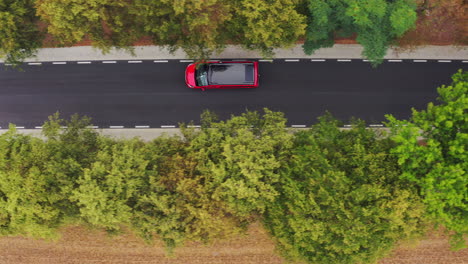 drone arriba abajo asimétrico seguimiento disparo de minivan camper rojo con techo negro con paneles solares conduciendo a lo largo de una estrecha carretera no marcada en el callejón de árboles