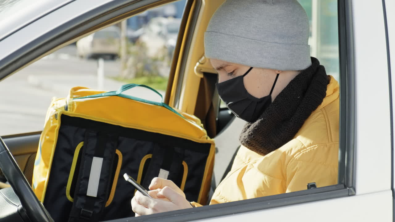 Courier in the car is on his phone, backpack for orders on the seat. Food delivery service