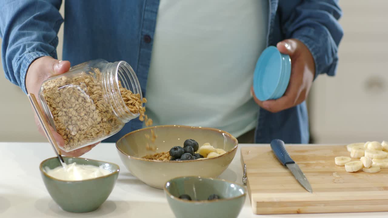 Uomo afroamericano che versa la granola da un barattolo in una ciotola dopo aver affettato le banane, tenendo un coperchio blu.