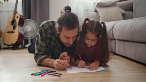 A-lonely-father,-a-brunette-man-in-a-green-checkered-shirt,-draws-with-his-little-daughter,-a-brunette-girl-in-a-pink-dress,-on-a-white-piece-of-paper-using-multi-colored-felt-tip-pens,-lying-on-the-floor-in-a-modern-apartment