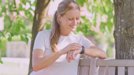 Frau-In-Fitnesskleidung-Sitzt-Auf-Einem-Sitz-Unter-Einem-Baum-Und-überprüft-Den-Aktivitätsmonitor-Auf-Der-Smartwatch