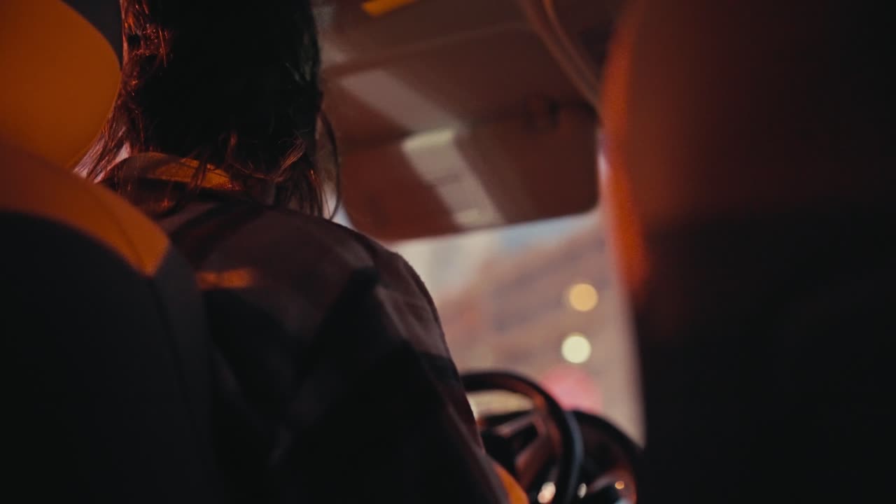 Rear view of a brunette man with long hair in a checkered shirt driving a car in the evening through the city after his work when he goes home. Evening ride around the city