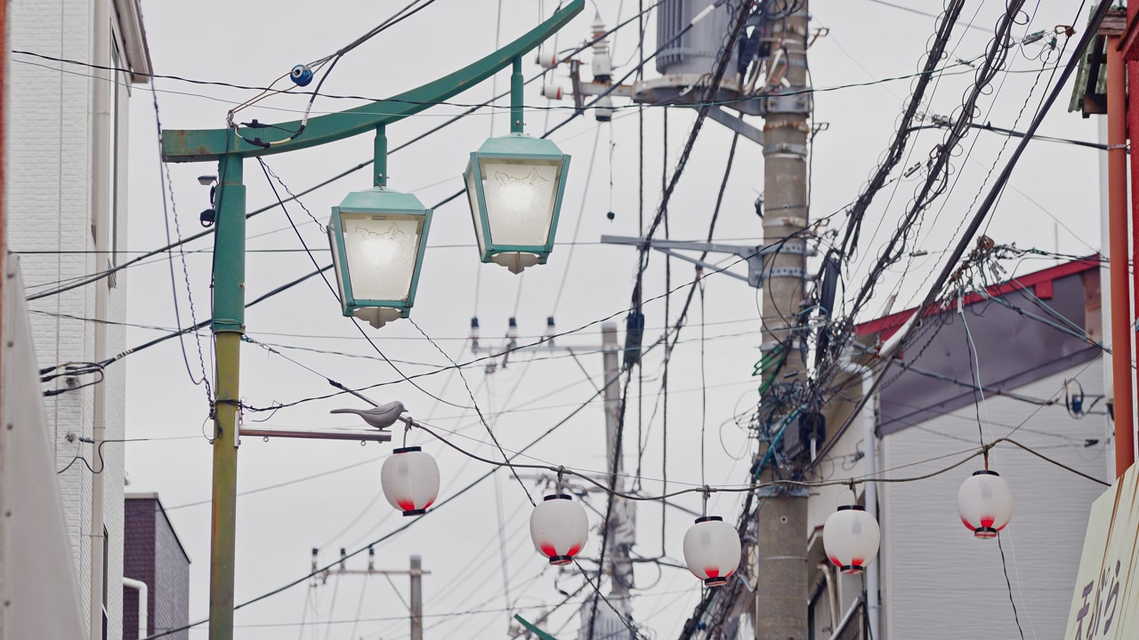 An intricate network of overhead power lines and cables frames traditional street lamps and white lanterns under an overcast urban sky.