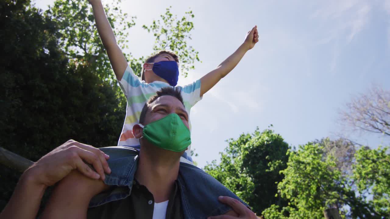 padre caucásico con máscara facial llevando a su hijo sobre sus hombros en el jardín