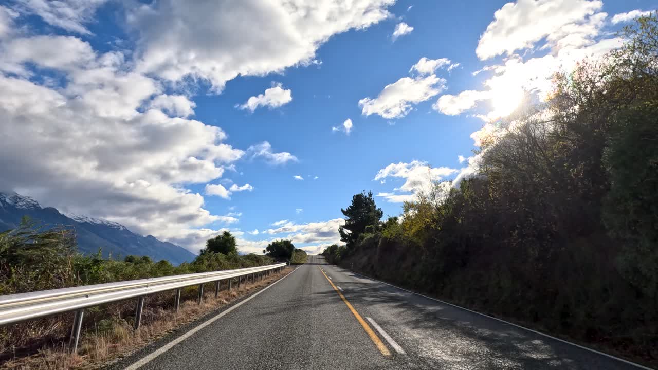 Vehicle travels sunlit mountain road with lake views, dynamic clouds, and lush roadside foliage