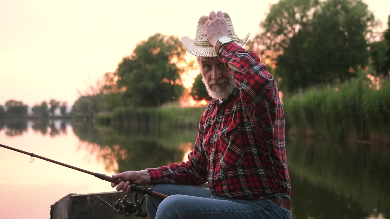 retrato de un pescador senior poniéndose sombrero y sentado en el muelle del lago mientras sostiene una caña de pescar al atardecer