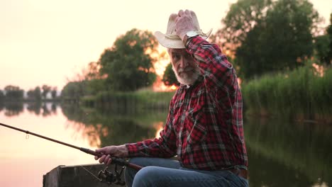 Portrait-of-a-senior-fisherman-putting-on-hat-and-sitting-on-the-lake-pier-while-holding-a-fishing-rod-at-sunset